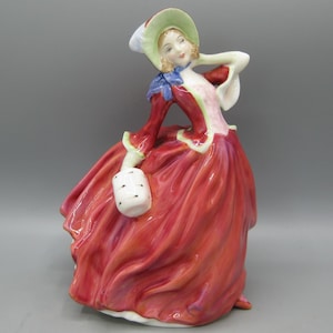 May include: A porcelain figurine of a person wearing a wide-brimmed light green hat, a blue neck scarf, and a long, flowing red dress with a pink bodice. The figure holds a small white cylindrical muff in one hand and has a hand raised to the hat.