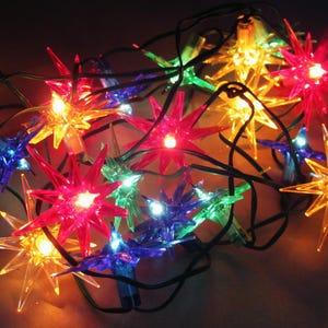 May include: A string of star-shaped Christmas lights in various colors, including red, yellow, blue, and green. The lights are strung on a black wire and are illuminated, creating a festive and colorful display.