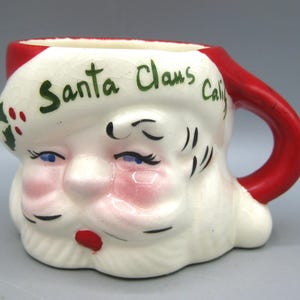 May include: A ceramic Santa Claus mug with a red handle and rim. The mug is white with a painted Santa face, including blue eyes, a red nose, and a white beard. The words "Santa Claus Calif" are written in green.