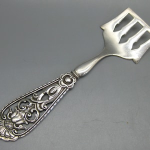 May include: Silver-toned serving utensil with a decorative handle and slotted head. The handle features an ornate floral design with a sunflower and scrolling details. The head has three slots. The utensil is likely for serving cheese or other foods.