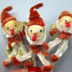 May include: Three vintage Christmas elf ornaments. Each elf has a tan face with blue eyes, a red nose and tongue, and a red and brown hat. The elves have white bodies with red and green accents.