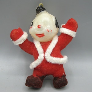May include: Vintage Christmas ornament in the shape of a cheerful figure. The figure is dressed in a red felt suit with white trim and matching mittens. The face is white with black hair and a black hat. A gold string is attached for hanging.