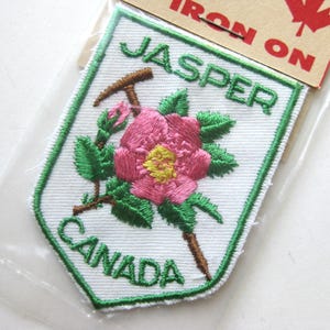 May include: Embroidered patch featuring a pink flower with a yellow center, green leaves, and a brown pickaxe. The patch reads "JASPER CANADA" in green lettering, outlined in white. The patch is on a white background with a green border.