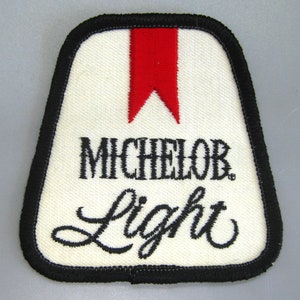 May include: A white, embroidered patch with a black border. The patch features the words "MICHELOB Light" in black script and a red vertical accent at the top. The patch has a textured appearance.