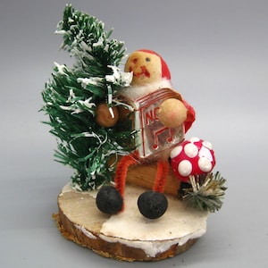 May include: A vintage Christmas decoration featuring a figure holding a green tree and a musical instrument. The figure has a red hat and orange pipe cleaner legs. A red and white mushroom and black ball feet complete the scene.