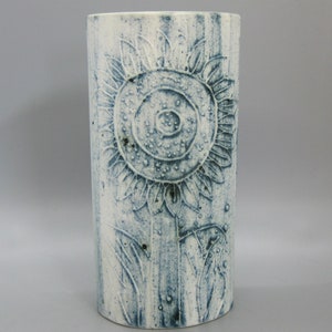 Puede incluir: Jarrón cilíndrico de cerámica con superficie texturizada blanquecina. Un gran diseño de girasol detallado está en relieve en la parte delantera, con acentos azul-gris. El jarrón tiene un patrón de rayas verticales y es adecuado para la decoración del hogar.