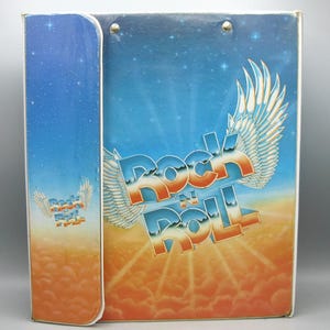 May include: A rectangular binder with a blue and orange gradient background featuring a starry sky and clouds. The text "Rock N Roll" is printed in large, stylized white and orange letters with white wings extending from the sides of the lettering.