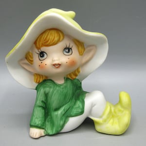 May include: A ceramic figurine of a seated elf with blonde hair, wearing a large yellow hat, a green tunic, and yellow pointed shoes. The elf has pointed ears, rosy cheeks, and freckles, with a smooth, glossy finish and painted facial features.