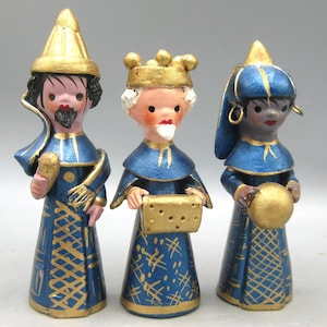 May include: Three ceramic figurines of the Three Wise Men. Each figure is dressed in blue robes with gold accents and holds a gold object. The figures have different skin tones and facial features, and wear gold crowns or hats. A festive holiday decoration.