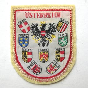 May include: Embroidered patch featuring the word "ÖSTERREICH" at the top. The patch has a shield shape with a yellow border and a red stitched edge. It displays the Austrian coat of arms and several regional crests.