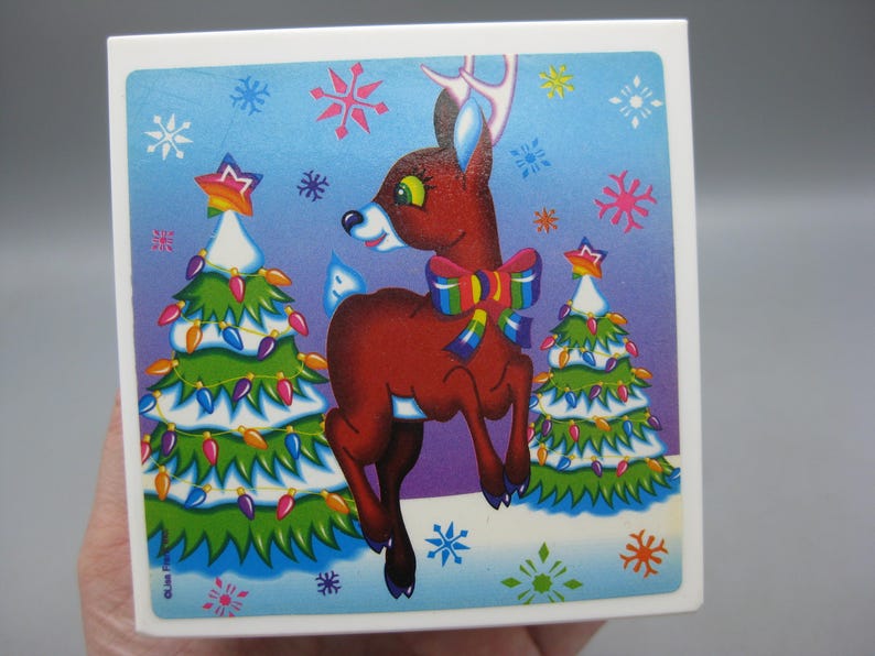 May include: A square Christmas-themed image featuring a cartoon reindeer with a colorful bow, standing between two decorated Christmas trees. The background is a gradient of blue and purple with snowflakes.