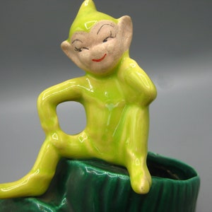 Pode incluir: Estatueta de cerâmica vintage de um elfo sentado, com corpo verde e rosto bege. O elfo está encostado numa base verde escura. O elfo tem um sorriso vermelho e olhos fechados. A estatueta é provavelmente um vaso.