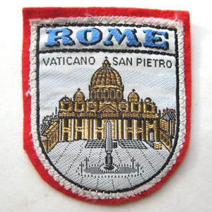 May include: A rectangular embroidered patch with a red felt border. The center features a white background with a gold-colored depiction of St. Peter's Basilica. The text ROME is in blue at the top, with VATICANO SAN PIETRO printed in black below.