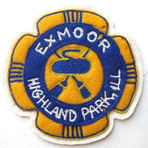 May include: A vintage felt patch in a four-leaf clover shape. The patch features a blue ring with the words "EXMOOR HIGHLAND PARK, ILL" embroidered in white. The center has a blue design on a yellow background.