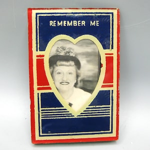 May include: Vintage rectangular photo frame with a heart-shaped opening. The frame is red, blue, and yellow, with the words "REMEMBER ME" at the top. The black and white photo features a woman wearing a hat.