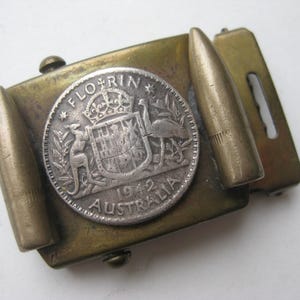 May include: Vintage brass belt buckle featuring a silver coin with an Australian coat of arms and the text "Florin 1942 Australia." The buckle has two bullet-shaped accents on either side of the coin.