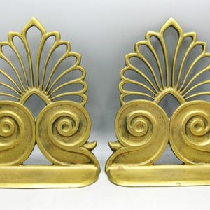 May include: A pair of antique gold-coloured bookends. Each bookend has a fan-shaped top and a pair of swirling scroll details. The bookends have a rectangular base and are made of metal. They are designed to hold books.
