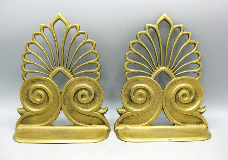May include: A pair of antique gold-coloured bookends. Each bookend has a fan-shaped top and a pair of swirling scroll details. The bookends have a rectangular base and are made of metal. They are designed to hold books.