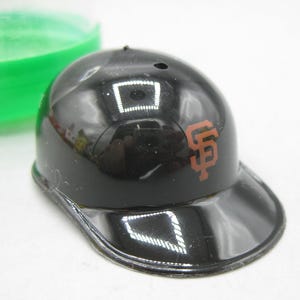 Op de afbeelding: Een kleine, zwarte honkbalhelm met een glanzende afwerking. Het logo van de San Francisco Giants staat in oranje op de zijkant. De helm heeft een gebogen klep. Op de achtergrond is een groene container te zien.