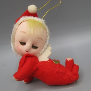 May include: A vintage Christmas ornament in the shape of a baby angel. The doll has a red outfit and hat, with a white halo and wings. The baby's face is made of fabric with closed eyes and blonde hair. A gold string is attached for hanging.