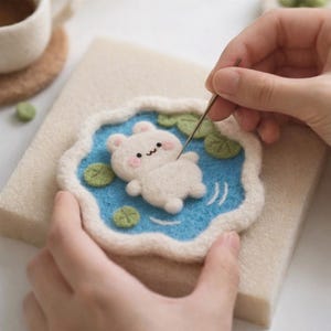 以下が含まれることがあります： 青い池に浮かぶ白いクマのフェルトクラフトプロジェクト。クマはピンクの頬と縫い付けられた笑顔をしています。池は白い縁で囲まれ、すべてベージュのフェルトベースにあります。針を使ってフェルトを加工しています。