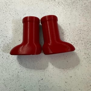 May include: Two small, bright red boots are displayed side-by-side. The boots have a rounded toe and a short, cylindrical shaft. The boots are made of a smooth material and cast a shadow on the light-colored surface.