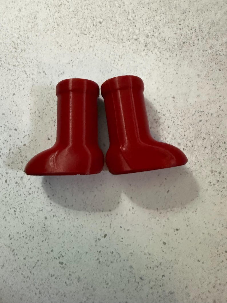 May include: Two small, bright red boots are displayed side-by-side. The boots have a rounded toe and a short, cylindrical shaft. The boots are made of a smooth material and cast a shadow on the light-colored surface.