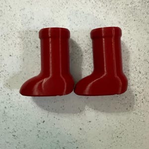 May include: Two small, bright red boots. The boots have a rounded toe and a cylindrical upper. The boots are likely decorative or for a small figurine. The boots are made of a solid material.