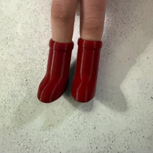 May include: A close-up of two doll legs wearing bright red boots. The boots are a solid, glossy red and have a simple, rounded design. The background is a speckled, light-colored surface.