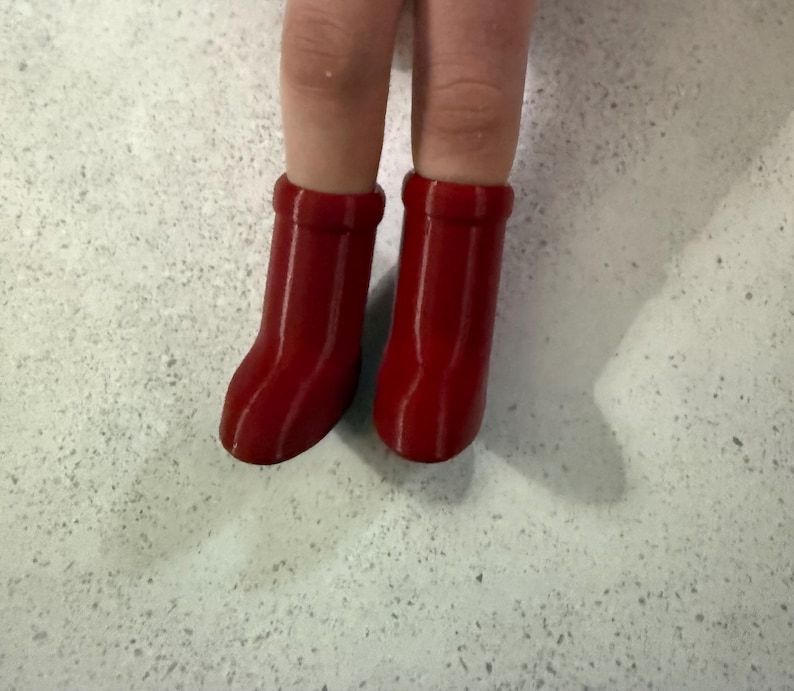 May include: A close-up of two doll legs wearing bright red boots. The boots are a solid, glossy red and have a simple, rounded design. The background is a speckled, light-colored surface.