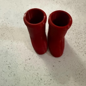 May include: Two bright red, cylindrical objects with a smooth, glossy finish. The objects have a flared top and a rounded base, suggesting a boot-like shape. They are positioned side-by-side against a speckled, light-colored surface.