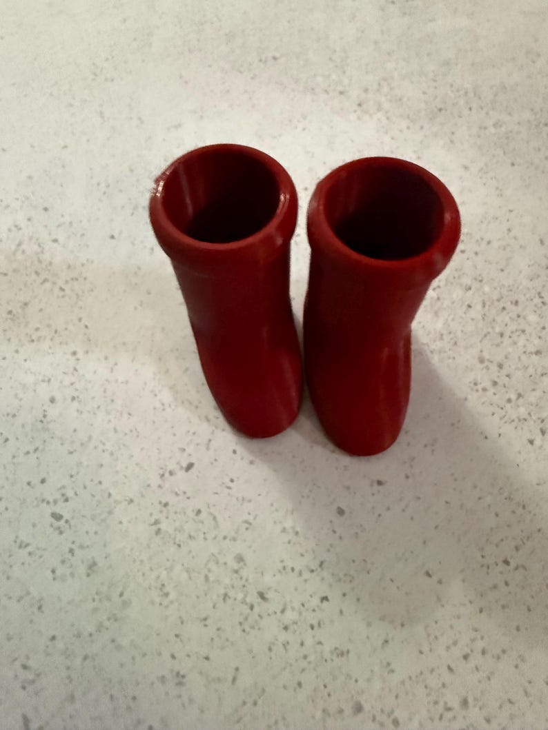 May include: Two bright red, cylindrical objects with a smooth, glossy finish. The objects have a flared top and a rounded base, suggesting a boot-like shape. They are positioned side-by-side against a speckled, light-colored surface.