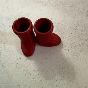 May include: Two small, bright red boots. The boots are made of a solid material and have a smooth finish. They are positioned on a light-colored surface.