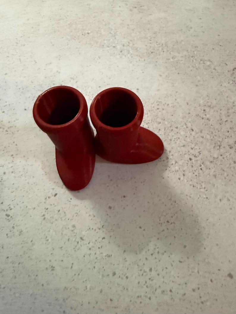May include: Two small, bright red boots. The boots are made of a solid material and have a smooth finish. They are positioned on a light-colored surface.