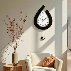 May include: A contemporary wall clock featuring a black, teardrop-shaped frame and a white clock face. The clock has black hands and a pendulum. The clock is mounted on a white wall. A vase with orange branches is on a wooden table.