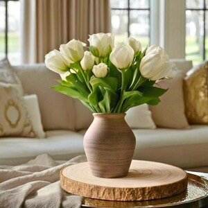 May include: A display of white tulips with green foliage in a brown ceramic vase. The vase is placed on a round wooden slab, set on a gold tray. A beige sofa and window are in the background.
