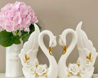 May include: Two white ceramic swan figurines with gold accents, forming a heart shape. Each swan has a gold beak and is adorned with white roses and gold leaf details. A vase of pink hydrangeas is in the background.