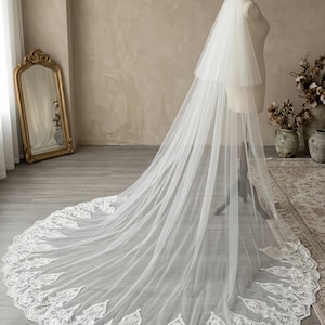 May include: A white bridal veil with a two-tiered design and a long train. The edge of the veil is adorned with a delicate, embroidered floral pattern. The veil is displayed on a mannequin in a well-lit room.
