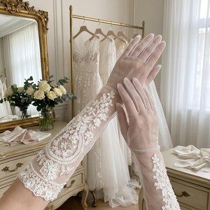 May include: Delicate, sheer white gloves with detailed floral lace patterns on the sleeves. The gloves are long, extending past the wrists, and are likely bridal wear. The background includes wedding dresses and a vintage mirror.