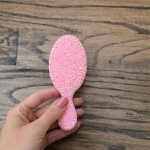Puede incluir: Cepillo para el cabello rosa, de forma ovalada, cubierto de pequeños pedrería brillantes. El cepillo tiene un mango y se sostiene sobre un fondo de madera. La pedrería le da al cepillo un aspecto texturizado.