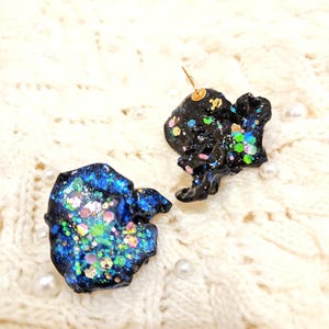 Könnte beinhalten: Zwei handgefertigte, abstrakte Ohrringe. Einer ist dunkelblau mit bunten Glitzer, der andere ist schwarz mit bunten Glitzer. Die Ohrringe liegen auf einer weißen, strukturierten Oberfläche mit verstreuten Perlen. Die Ohrringe haben goldfarbene Stifte.