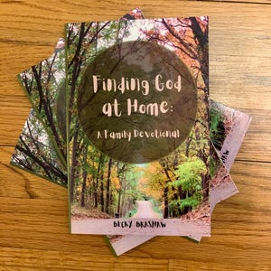 May include: Several copies of a book titled "Finding God at Home: A Family Devotional" by Becky Brashaw. The cover shows a forest road scene within a circular frame. The books are stacked on a wooden surface.