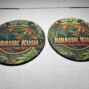 May include: Two circular coasters with a green and orange design. They feature a T-Rex, cannabis leaves, and the text "JURASSIC KUSH" and "KEEP IT LIT". The background includes a jeep and other foliage.