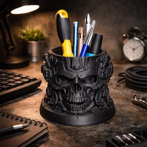May include: A black skull-shaped desk tidy holding pens, a screwdriver, and other stationery. The organiser has a detailed, textured design and is on a desk with other items in the background.