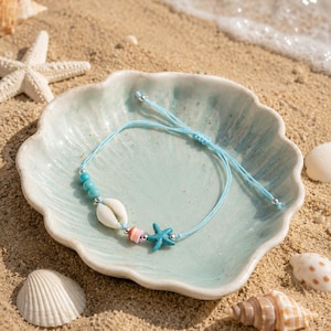 May include: A light blue cord bracelet featuring three small turquoise-colored beads, a white cowrie shell, a small striped cylindrical bead, and a blue starfish charm. The bracelet has a sliding knot closure with silver-toned metal end beads.