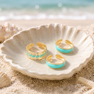 May include: Three gold-toned open rings featuring bright turquoise enamel accents. One ring displays a repeating leaf-like pattern, while the other two rings have a smooth, solid turquoise band design. Each ring has a polished metallic finish.