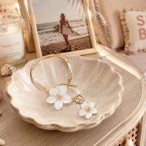 May include: A gold-toned bracelet featuring two white flower charms with yellow centers, designed with a toggle clasp. The bracelet rests inside a scalloped, shell-shaped ceramic dish with a metallic gold rim, showcasing a delicate and reflective finish.