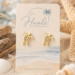 May include: A pair of gold-toned sea turtle stud earrings displayed on a rectangular card. The card features the text Haolé SURFBOARD CO. with a palm tree graphic and the text BEACHY LITTLE THINGS at the bottom, set against a beach scene.