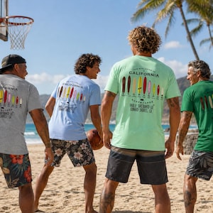 May include: Four people on a beach wearing t-shirts with surfboard graphics. The shirts are gray, light blue, green, and dark green. The text on the shirts reads "The Evolution of California Surfboards". The people are wearing floral print shorts.