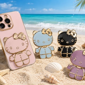 May include: A light pink smartphone case featuring a gold-outlined Hello Kitty character, accompanied by three separate Hello Kitty phone grips in light blue, black, and purple, each with gold-tone metal detailing and a bow on the head.